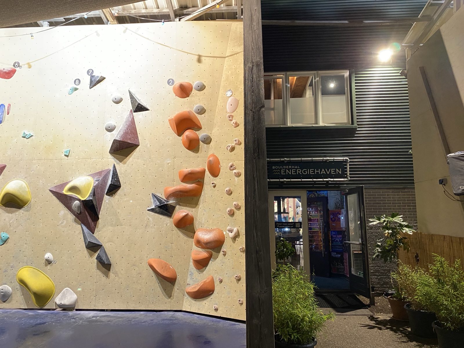 The bouldering wall of Boulderhal Energiehaven in the evening, with orange, yellow and grey holds on a light wooden wall, next to the open entrance under a sign with the hall's name.