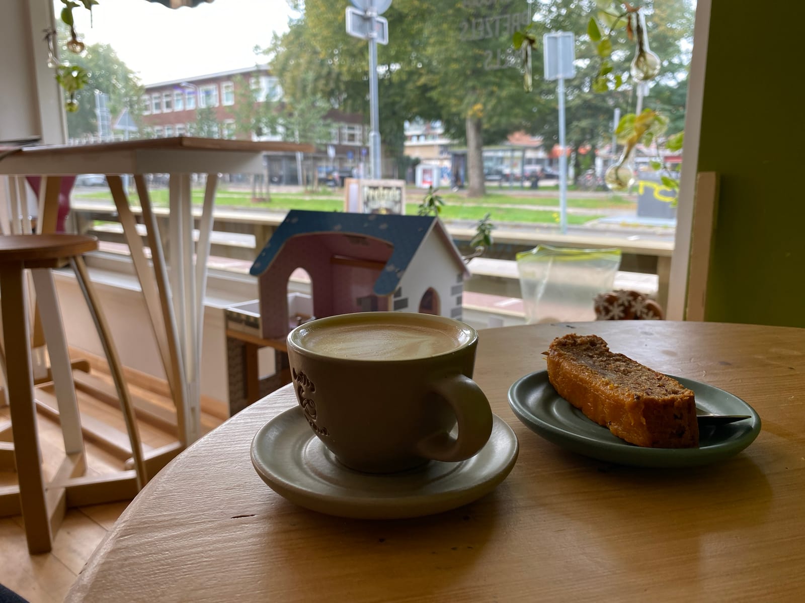 A cup of coffee and a slice of cake on a wooden table by a café window, with a toy dolls' house on the windowsill and a tree-lined street outside.