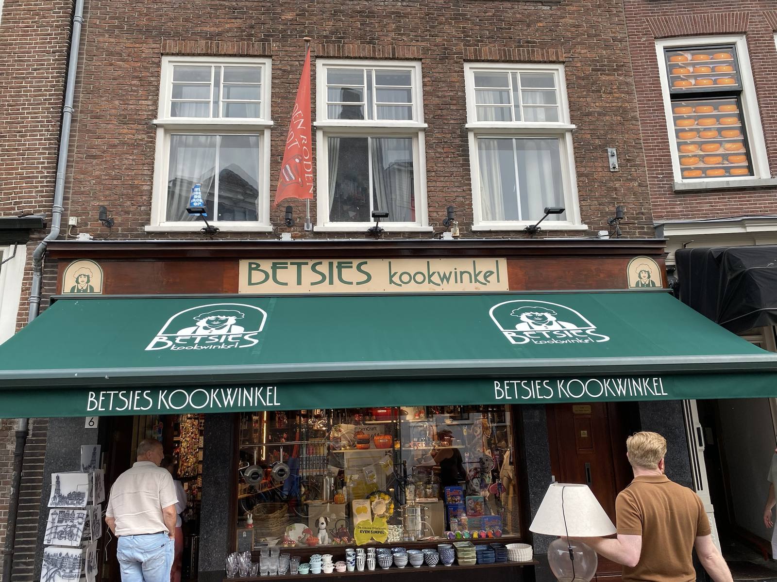 The front of Betsies Kookwinkel, a cooking shop in an old brick building, with its name on a green awning and a shop window full of pans and kitchenware. Two passers-by walk past, one of them carrying a lamp.