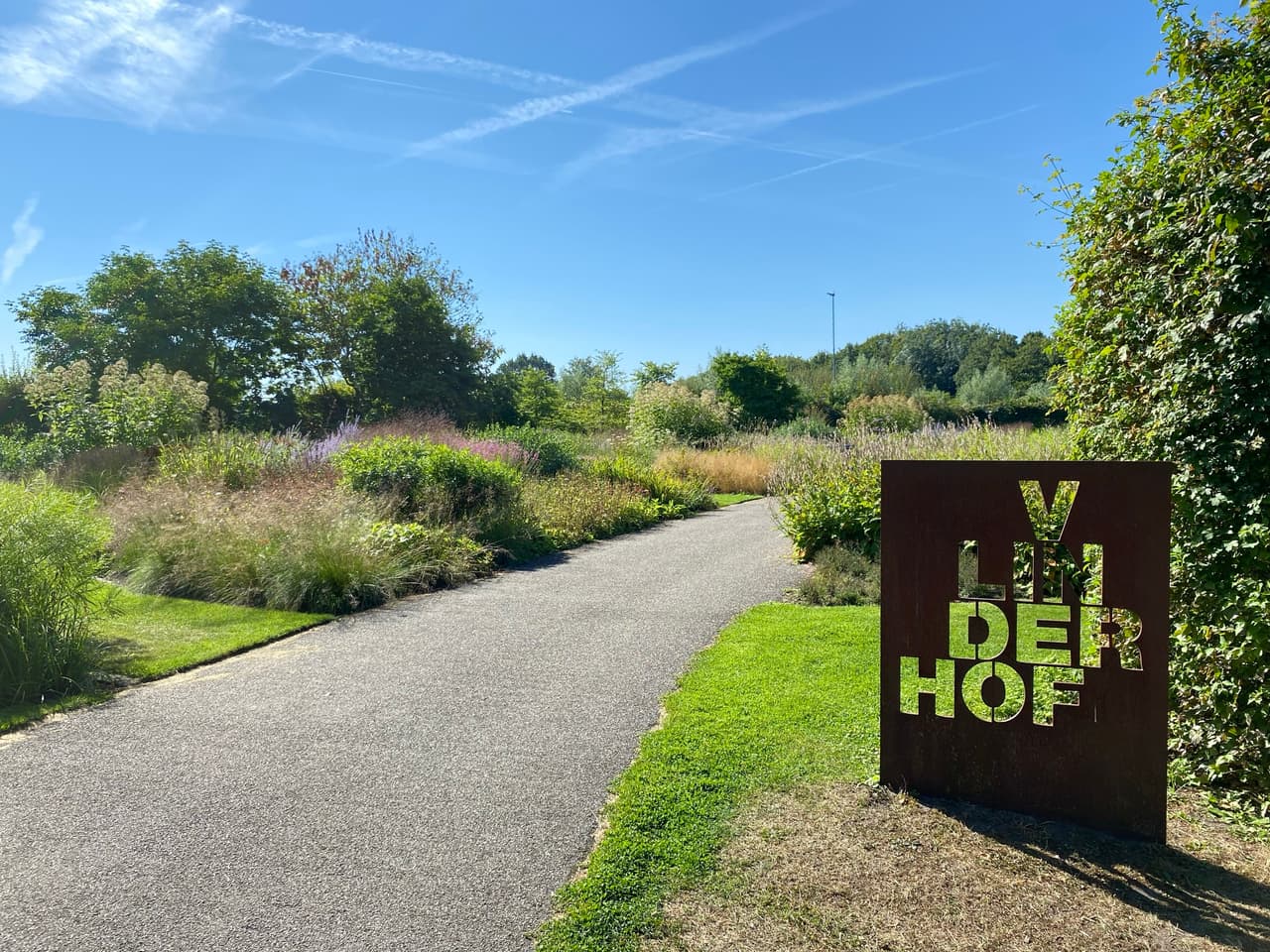 A gravel path curving through tall grasses and flowering perennials, with a rusted steel sign reading Vlinderhof standing in the grass on the right.